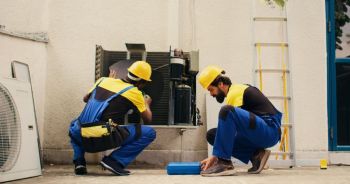 Two construction workers in hard hats crouching beside an outdoor electrical unit near a building wall